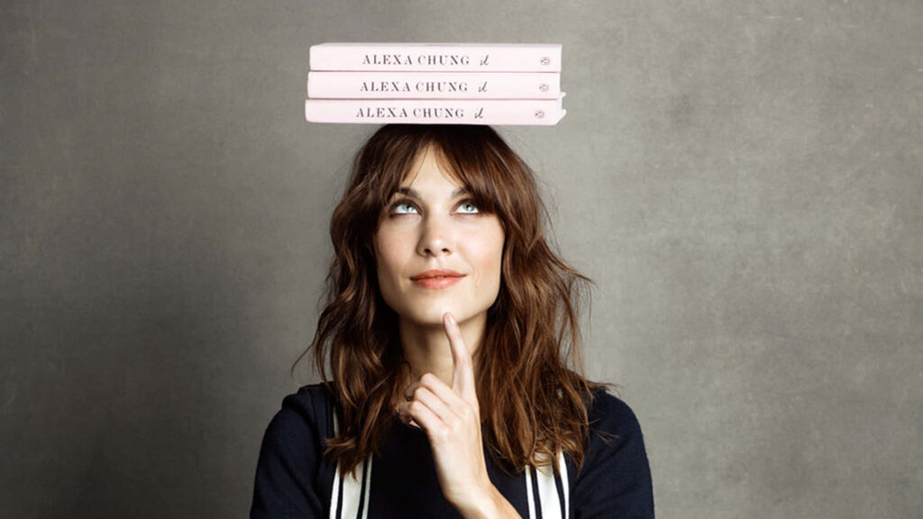 Alexa Chung thoughtful pose with finger on chin balancing three pink books on head wallpaper