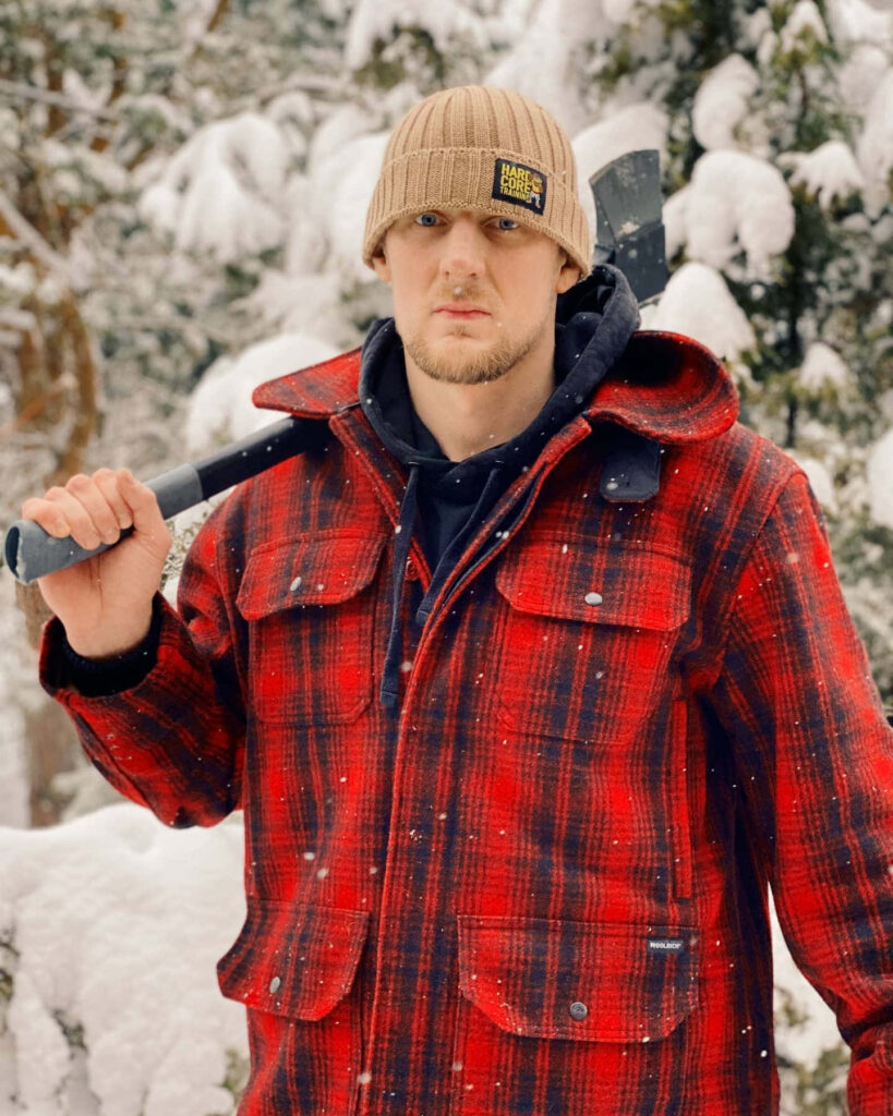 Alexander Volkov in red plaid jacket holding axe in snowy forest rugged winter outdoors scene Wallpaper
