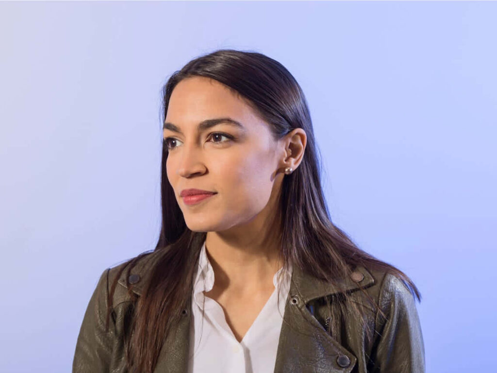 Alexandria Ocasio-Cortez in white shirt and green jacket standing confidently on blue background wallpaper