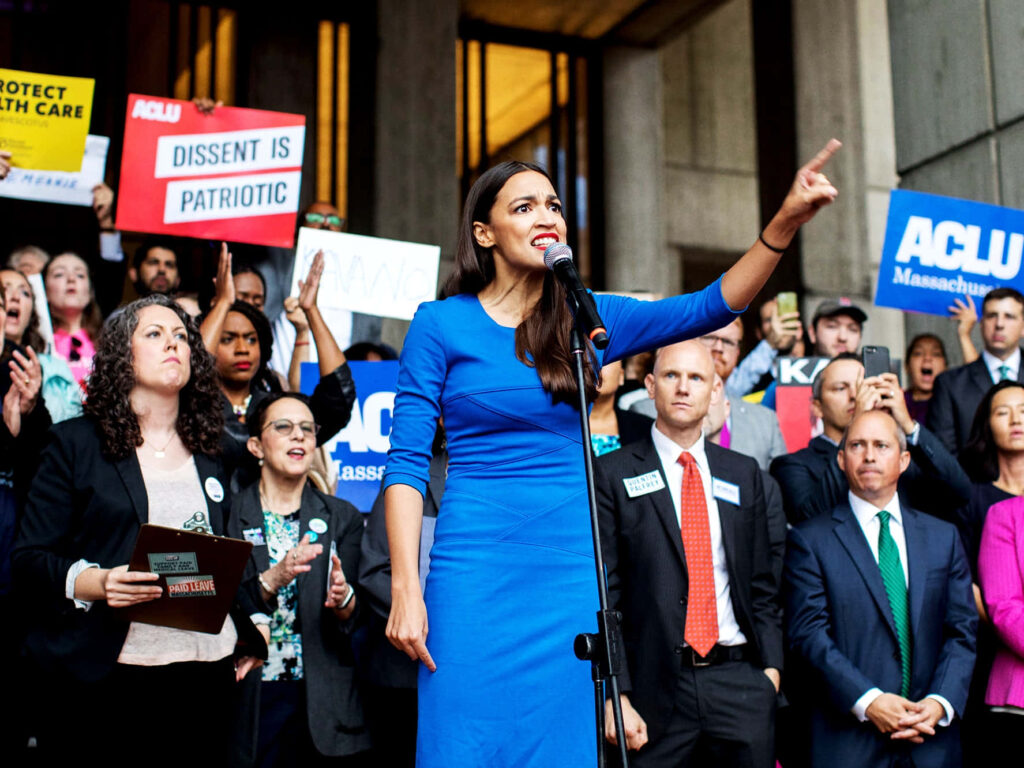 Alexandria Ocasio-Cortez speaking at rally in blue dress with supporters holding signs behind her Wallpaper