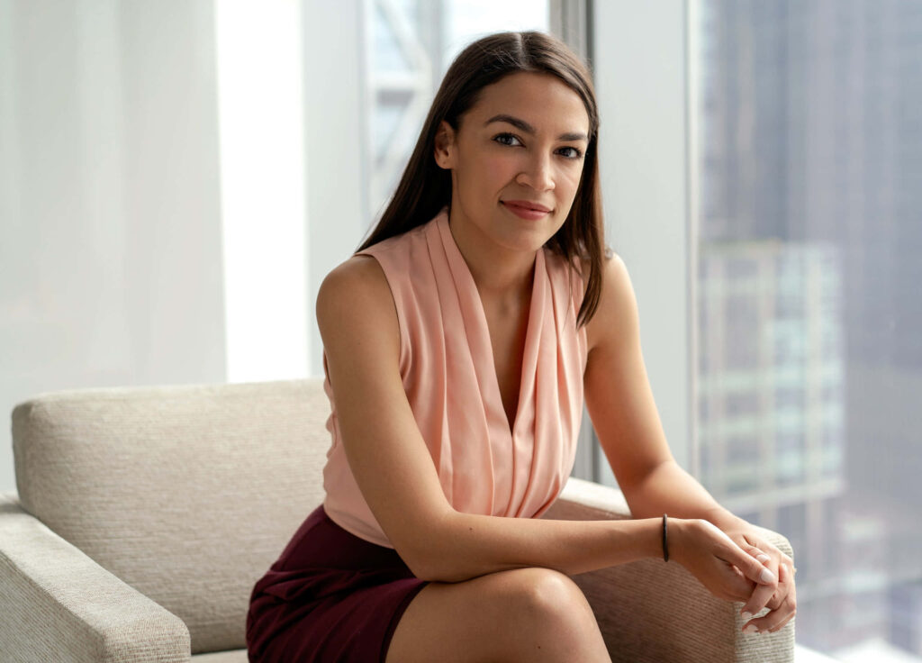 Alexandria Ocasio-Cortez seated by city window in blush top and burgundy skirt professional wallpaper