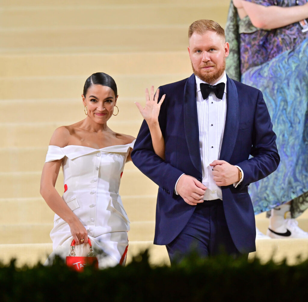 Alexandria Ocasio-Cortez and Riley Roberts in elegant formal attire posing on grand staircase wallpaper
