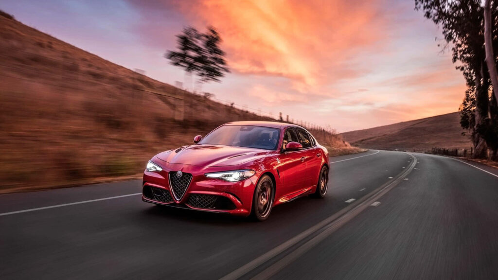 Alfa Romeo Giulia speeding on sunset road with vibrant orange sky and rolling hills background Wallpaper
