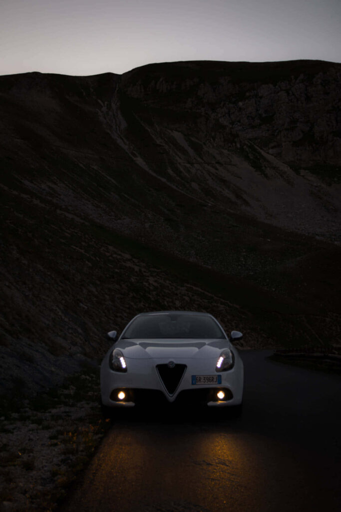 Red Alfa Romeo Giulietta with headlights on at dusk against dramatic mountain wallpaper background