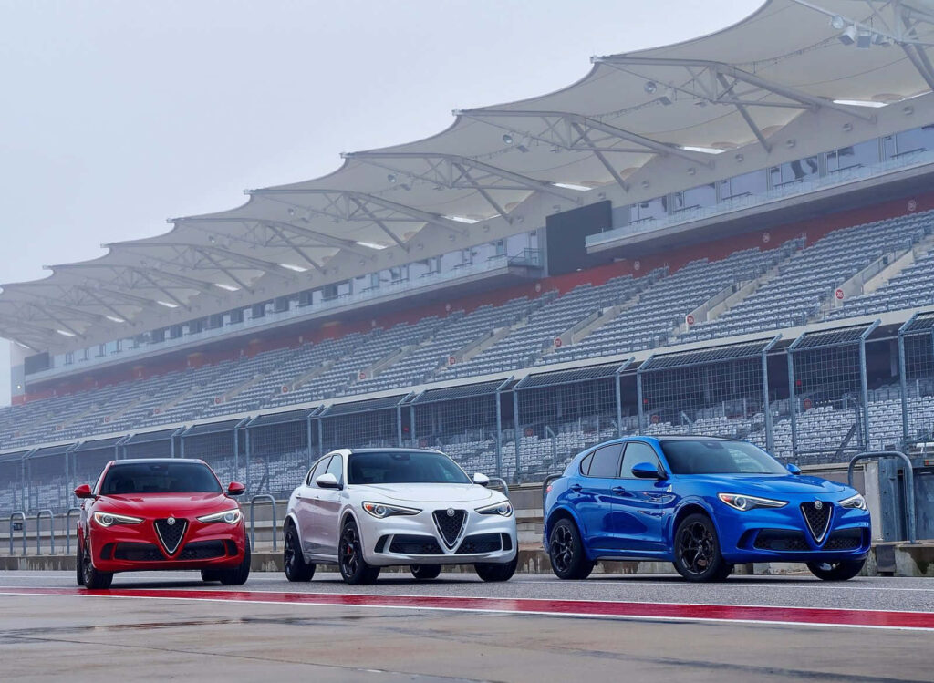 Alfa Romeo Stelvio SUVs in red white and blue lined up on racetrack with grandstands background Wallpaper