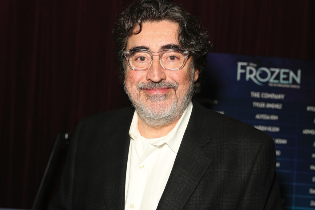 Actor Alfred Molina smiling at Frozen movie event with promotional backdrop wallpaper background