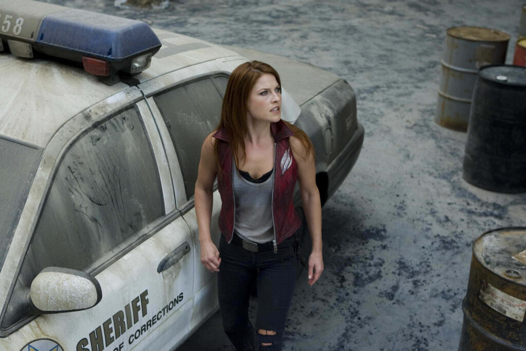Ali Larter in rugged outfit stands alert by weathered sheriff car in gritty abandoned setting wallpaper