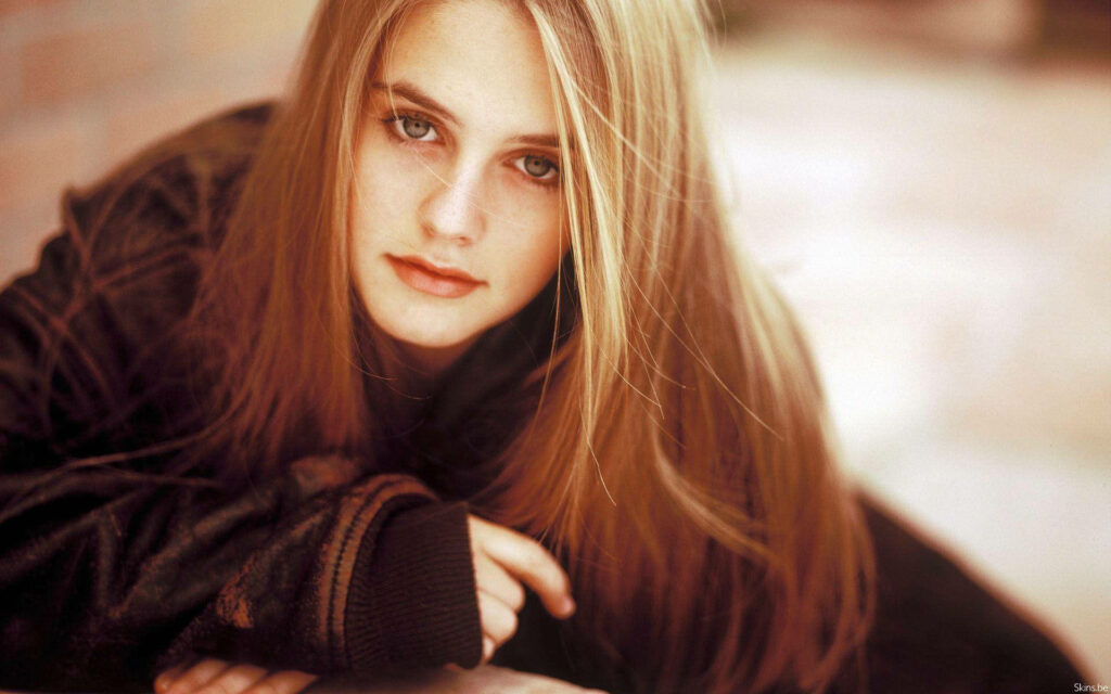 Alicia Silverstone wearing jacket with natural hair and warm intimate gaze against wallpaper background