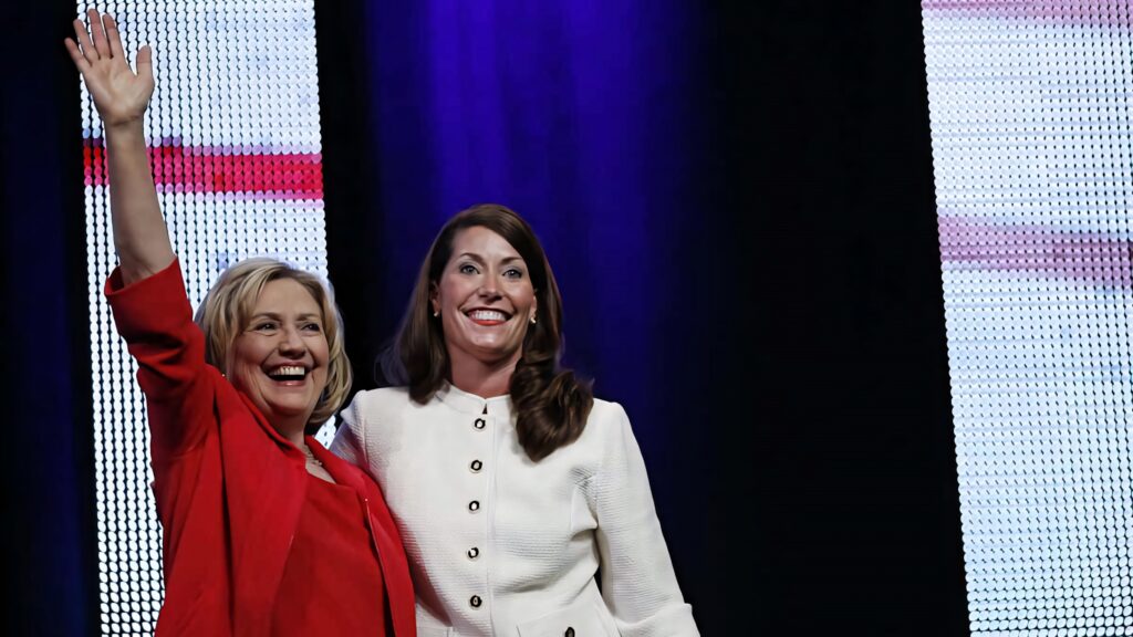 Alison Lundergan Grimes smiling and waving confidently on stage with vibrant wallpaper background