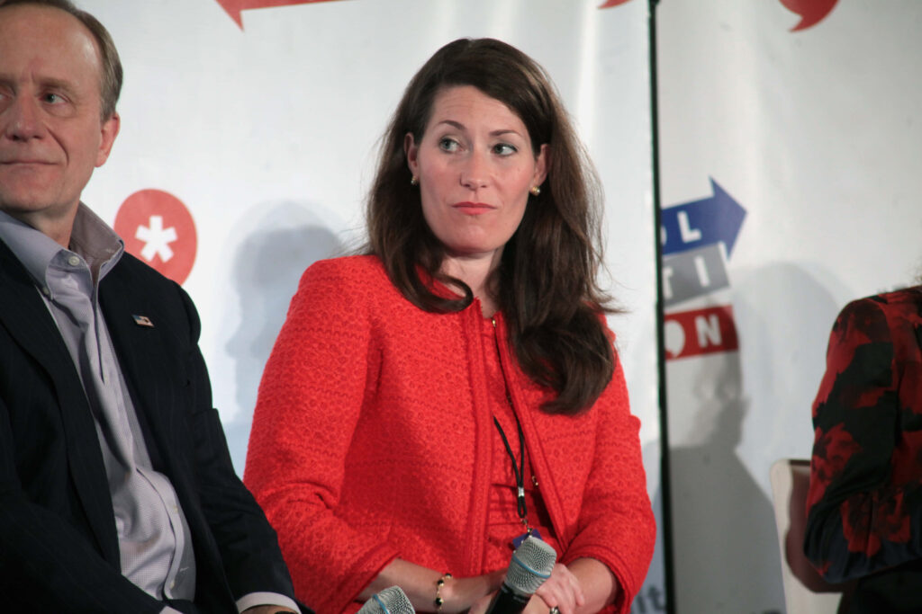 Alison Lundergan Grimes in red jacket holding microphone at conference with event backdrop Wallpaper