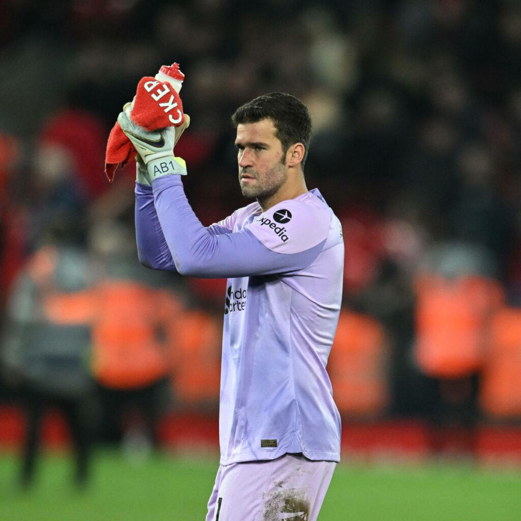 Alisson Becker goalkeeper holding water bottle and red towel on pitch with blurred stadium crowd background Wallpaper