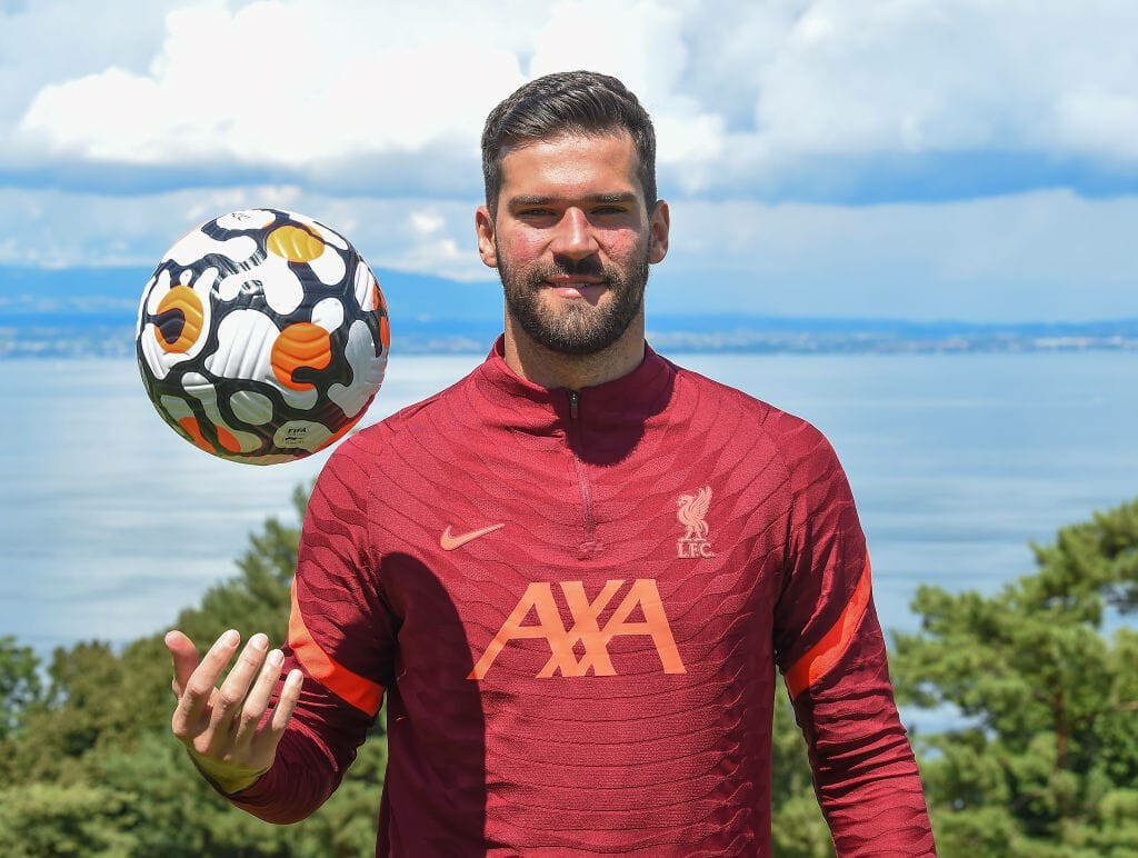 Alisson Becker in Liverpool FC training kit skillfully balancing football outdoors by scenic lake Wallpaper