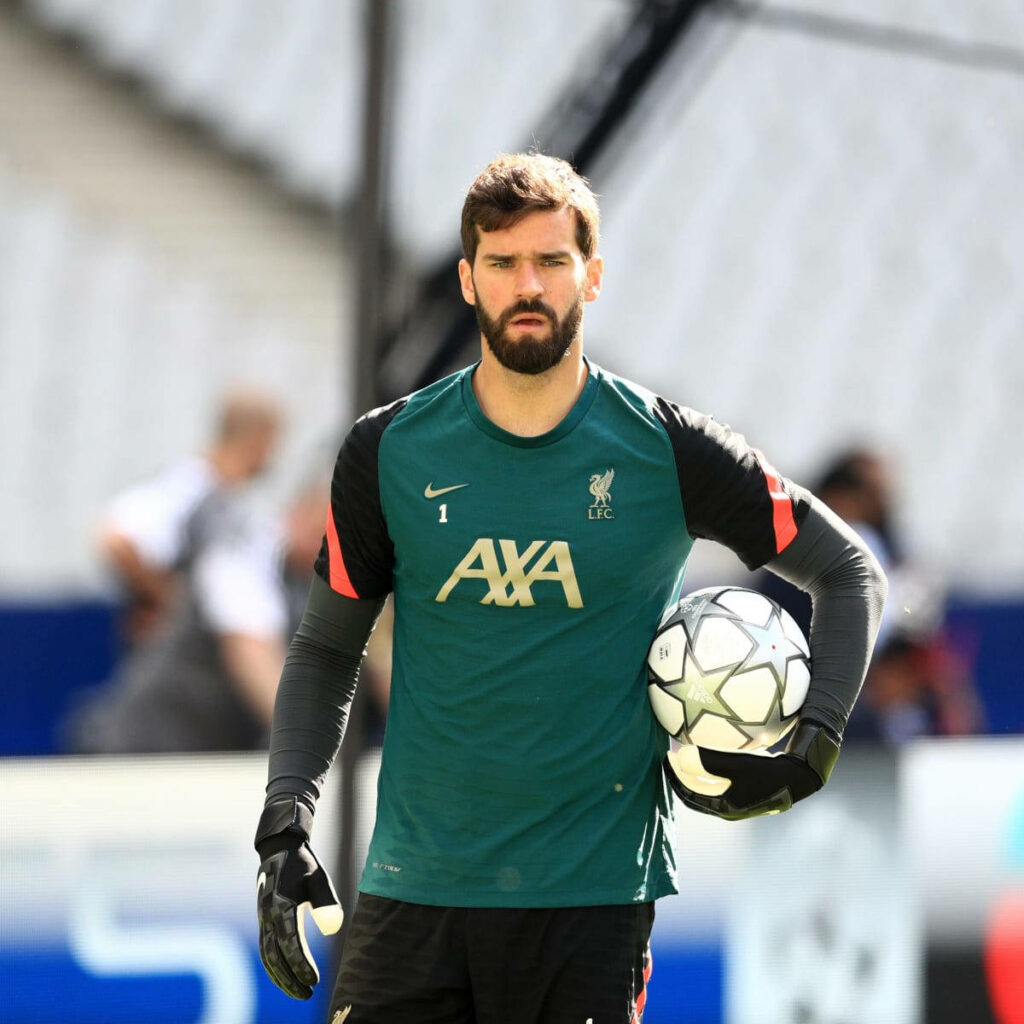 Alisson Becker in Liverpool training kit confidently holding football on field wallpaper background