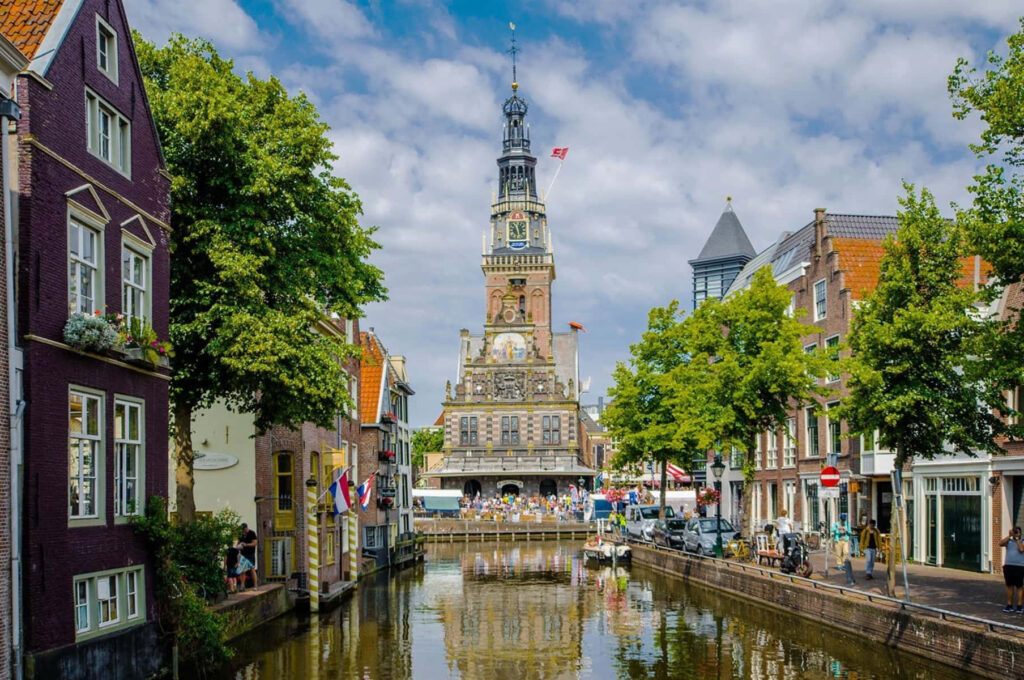 Scenic Alkmaar canal with historic buildings and landmark tower reflected in calm water under clouds Wallpaper