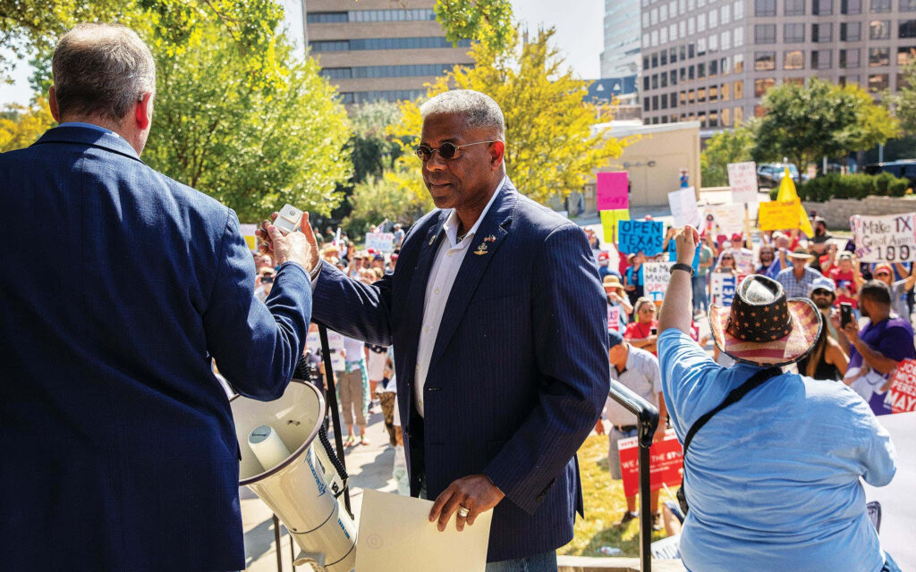 Allen West speaking at outdoor political rally engaging supporters with city buildings backdrop Wallpaper