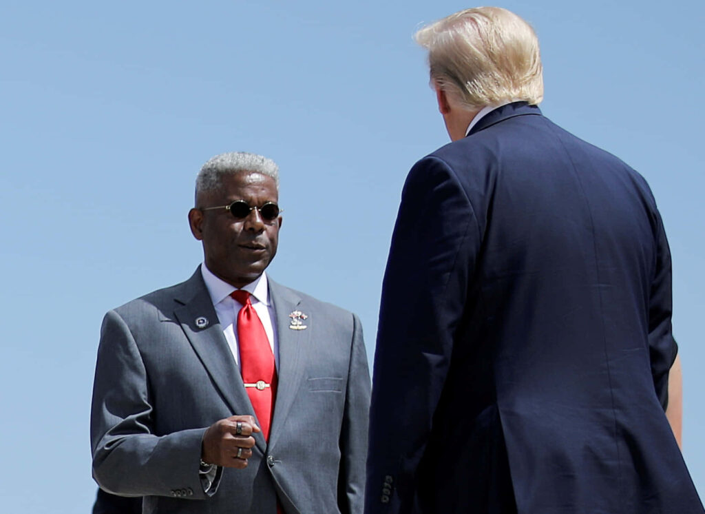 Allen West and Donald Trump in formal attire outdoors engaging in conversation under clear sky Wallpaper