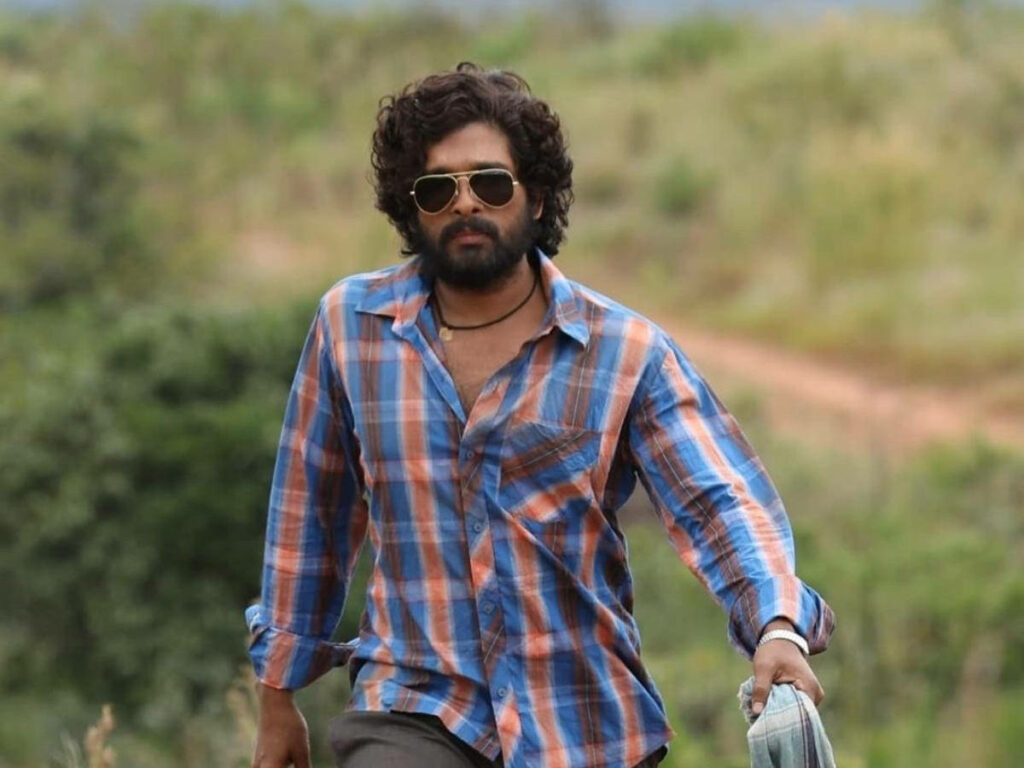 Allu Arjun as Puspa walking confidently on dirt path in field wearing checked shirt and sunglasses Wallpaper