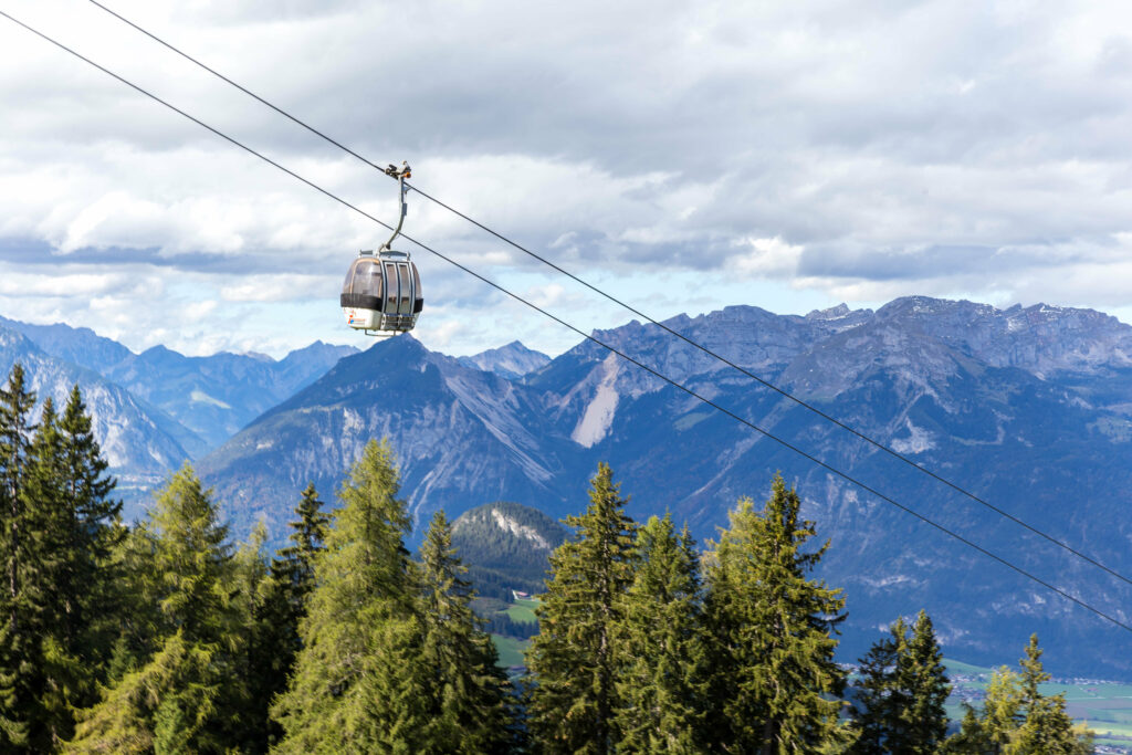 Cable car gliding over dense spruce forest with majestic alpine mountains under sky Wallpaper