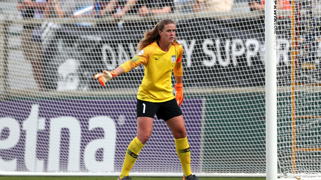 Alyssa Naeher focused in goalkeeper uniform ready for intense soccer match with goal net behind Wallpaper