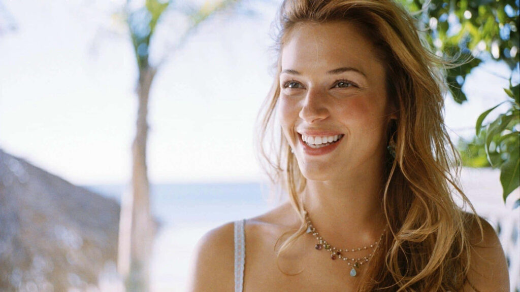 Amanda Righetti smiling brightly on sunny beach with lush greenery and distant ocean background Wallpaper