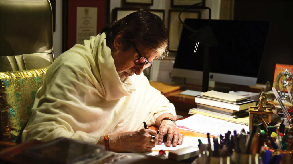 Amitabh Bachchan writing at desk in warm study surrounded by books and memorabilia wallpaper