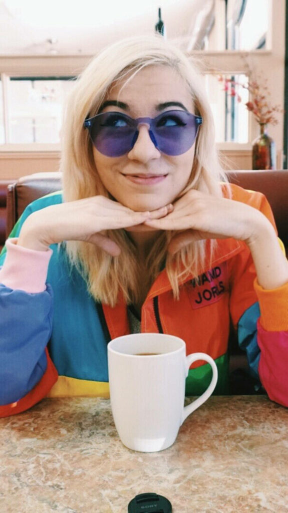 Amy Nelson in vibrant colorful jacket purple sunglasses sitting at table with white mug cheerful vibe Wallpaper