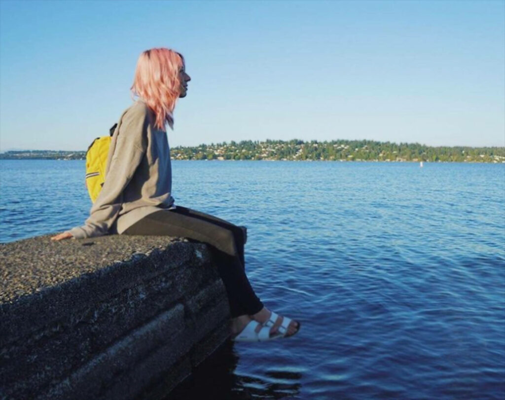 Amy Nelson sitting on concrete ledge overlooking calm blue waters and scenic Seattle skyline under clear sky Wallpaper