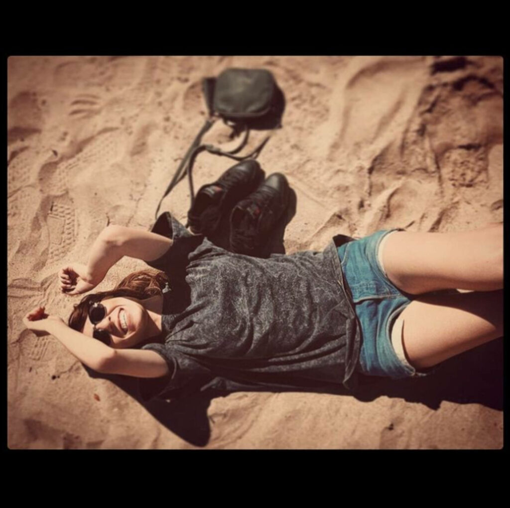 Amy Nelson relaxing on sunlit beach sand with backpack and shoes in casual laid-back scene Wallpaper