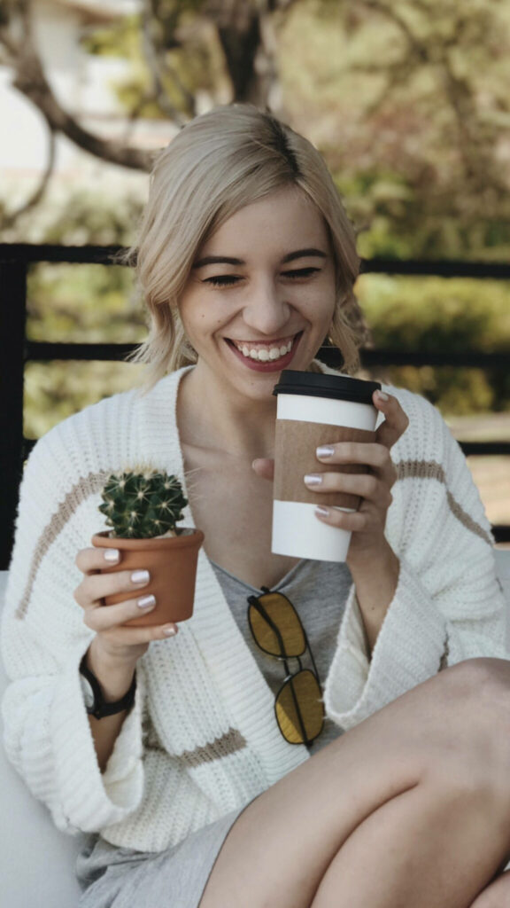 Amy Nelson smiling outdoors with coffee holding small cactus cozy white sweater cheerful vibe Wallpaper