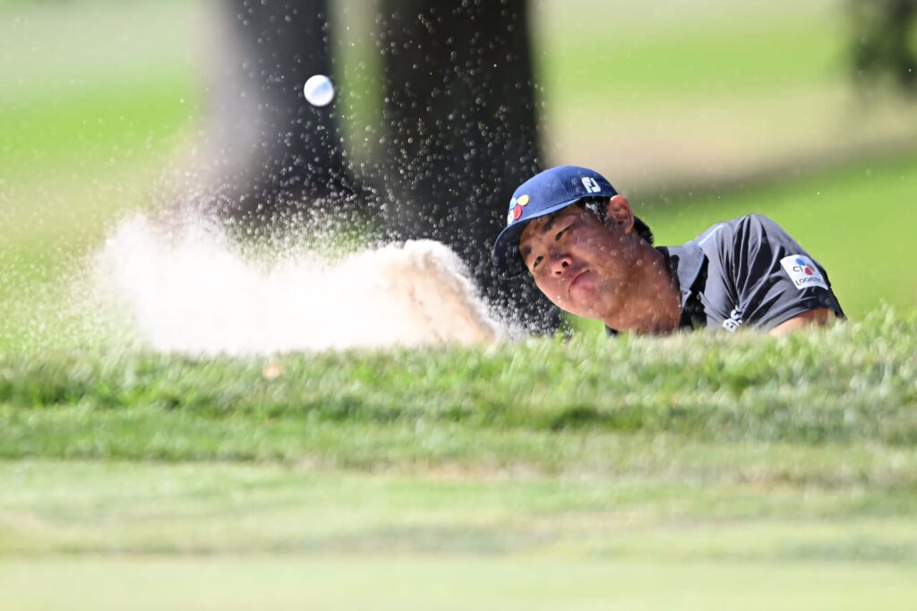 An Byeong-hun escaping sand trap with golf ball in motion on lush sunlit green golf course background Wallpaper