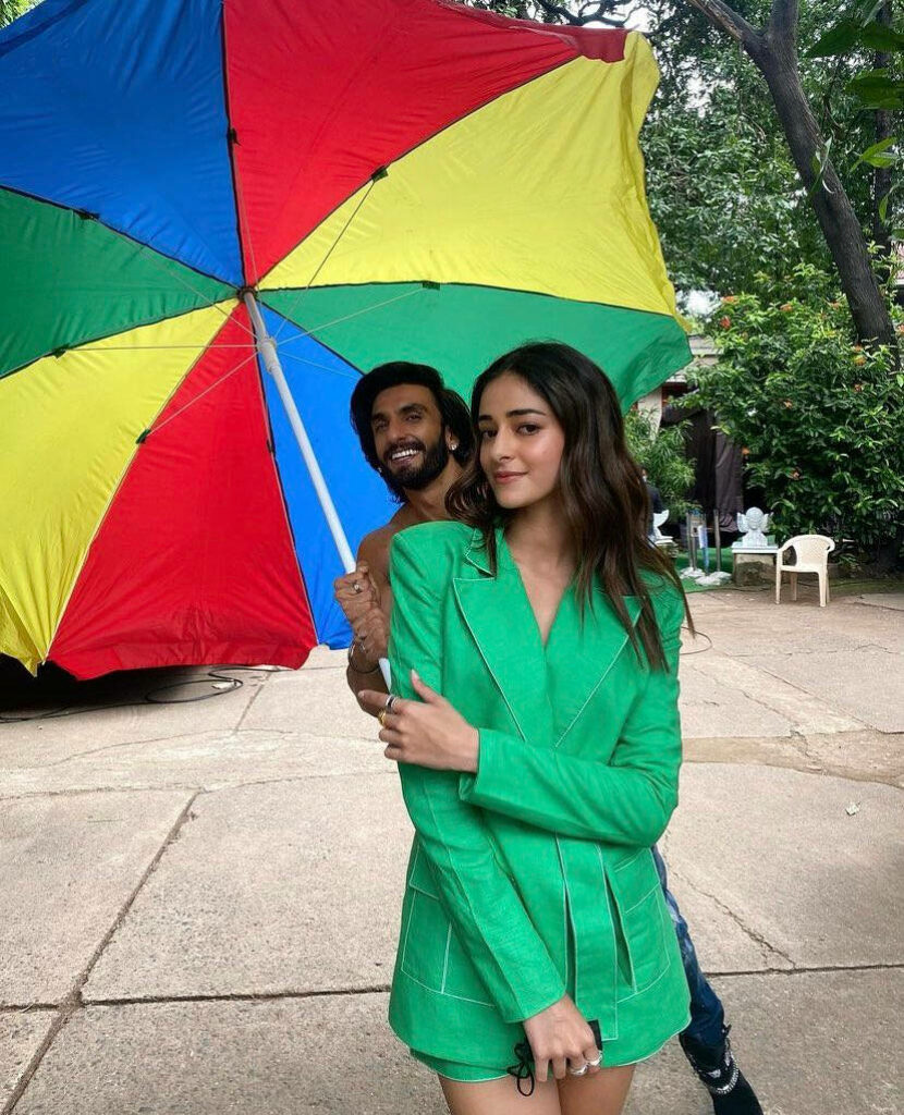 Ananya Pandey and Ranveer Singh smiling under colorful umbrella with green foliage background Wallpaper