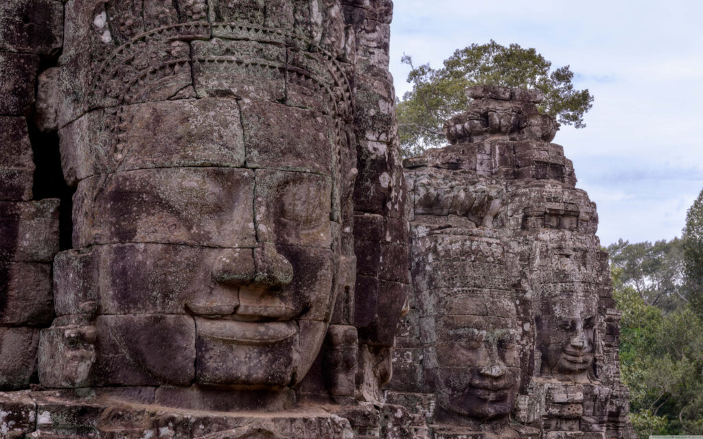 Ancient stone face statues of Angkor Thom with intricate carvings in lush green surroundings wallpaper