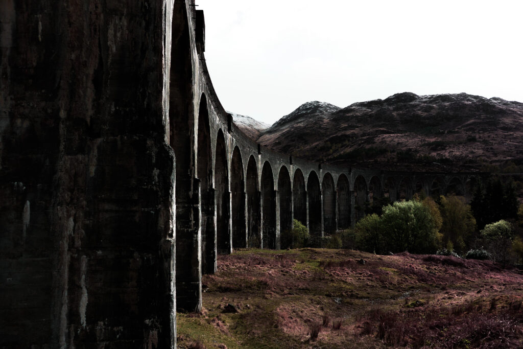 Ancient stone bridge with tall arches over wild landscape toward rugged mountains under pale sky Wallpaper