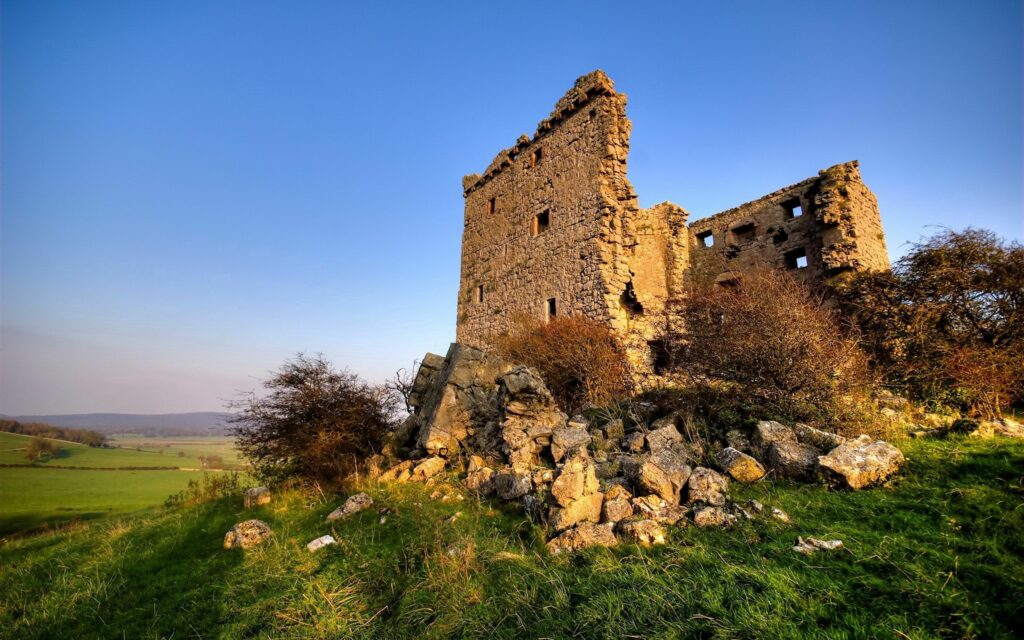 Weathered stone ruin in green grass bathed in golden sunlight under clear blue sky countryside Wallpaper