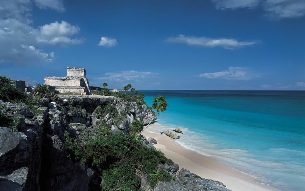 Ancient stone structure on rocky cliffs with palm trees overlooking blue water and sandy beach Wallpaper