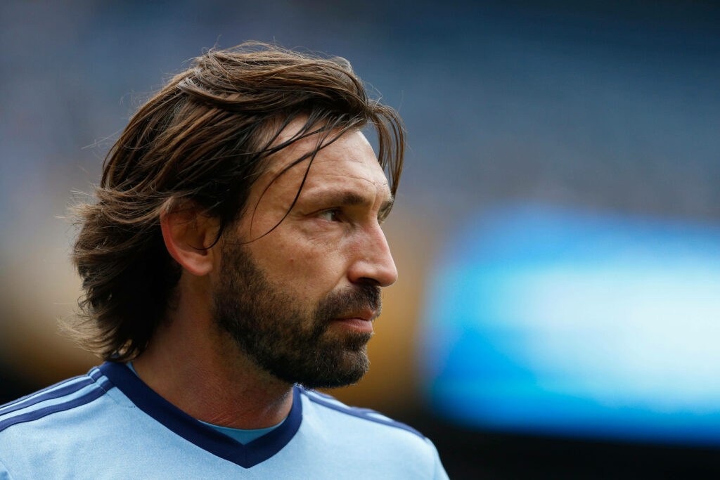 Andrea Pirlo headshot with thoughtful expression and signature hairstyle in blurred stadium background Wallpaper