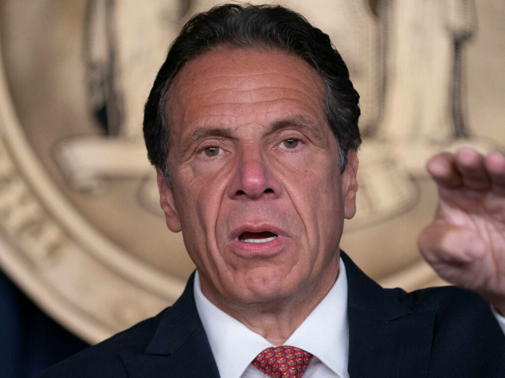 Andrew Cuomo tired and serious at press conference with large emblem wallpaper background