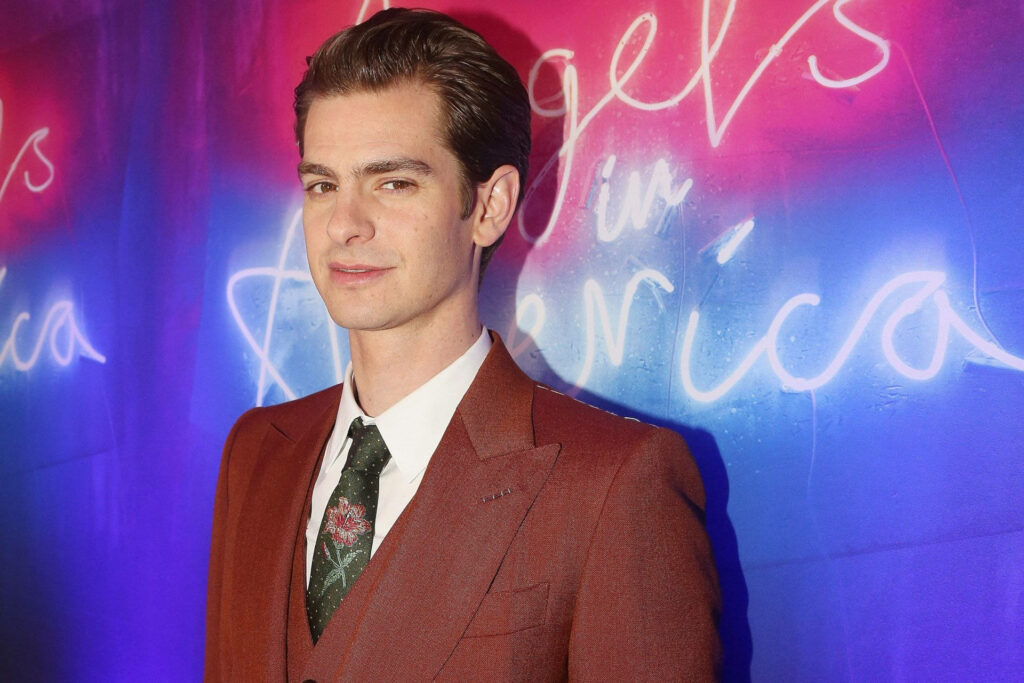 Andrew Garfield in brown suit with floral tie standing before vibrant Angels In America backdrop wallpaper