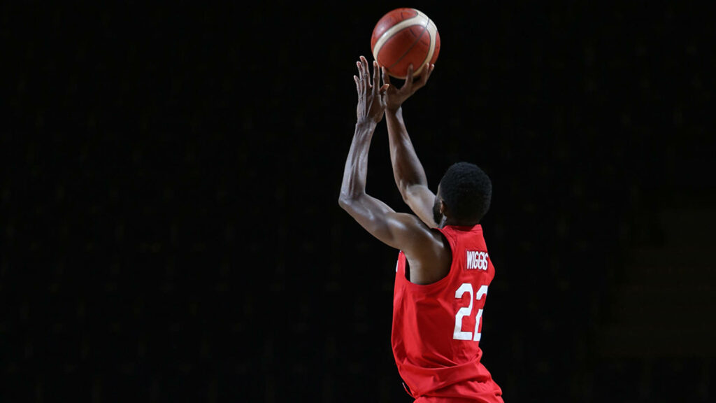 Andrew Wiggins mid-air shooting three-point shot in red jersey on dark basketball background Wallpaper