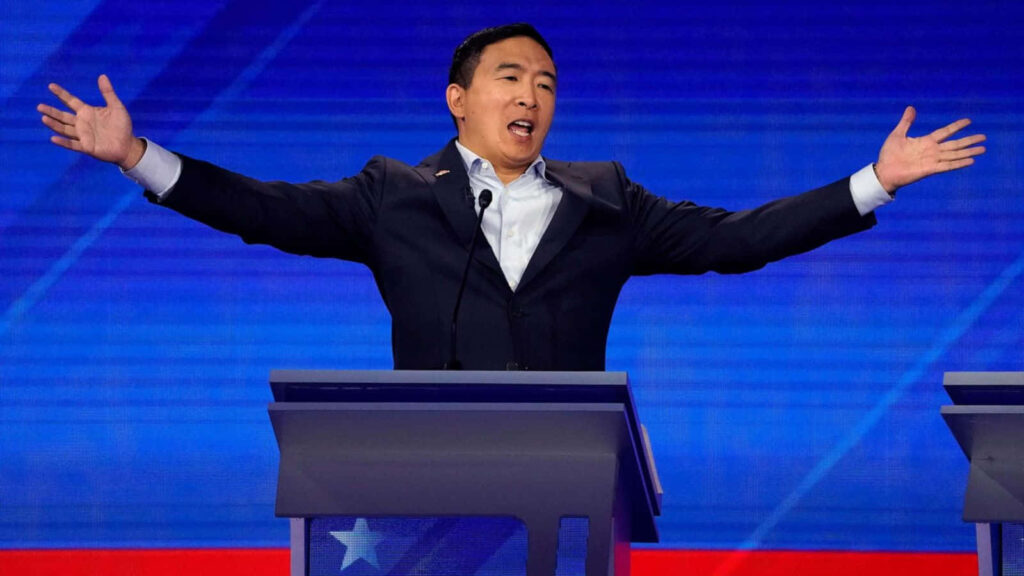Andrew Yang confidently speaking at debate podium with outstretched arms and vibrant blue background Wallpaper