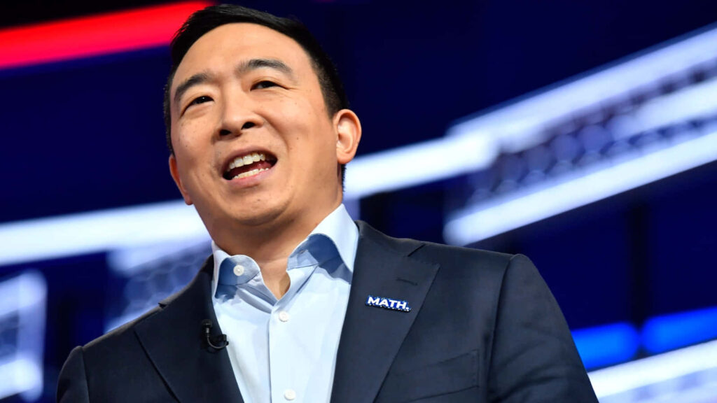 Andrew Yang speaking on stage in dark suit with MATH pin vibrant modern backdrop wallpaper