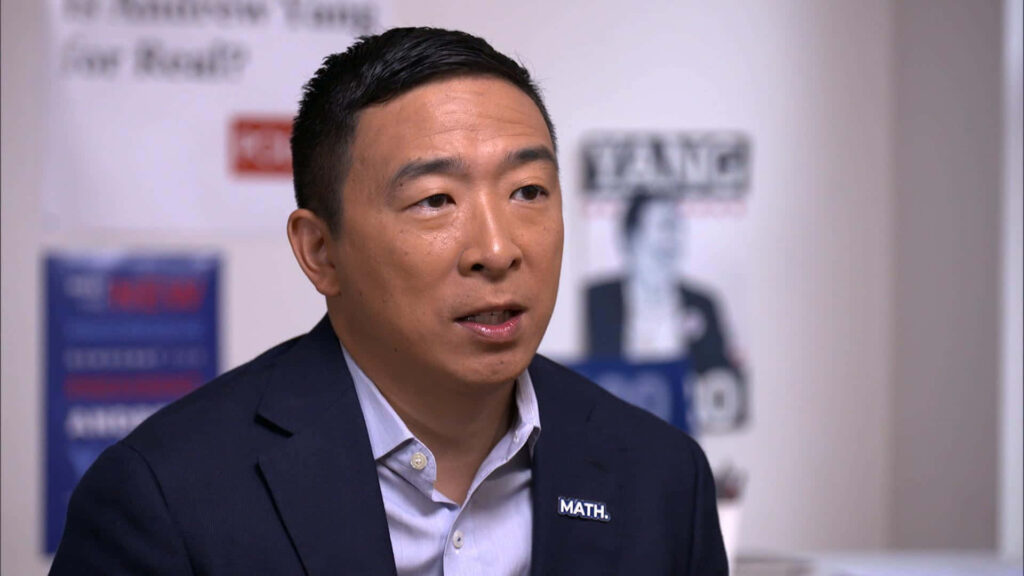 Close-up of Andrew Yang in dark blazer with Math pin and blurred campaign background wallpaper