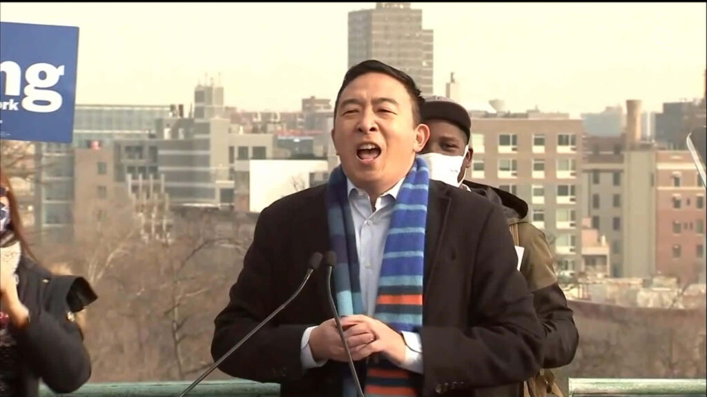 Andrew Yang speaking outdoors wearing colorful striped scarf dark coat city backdrop campaign event Wallpaper