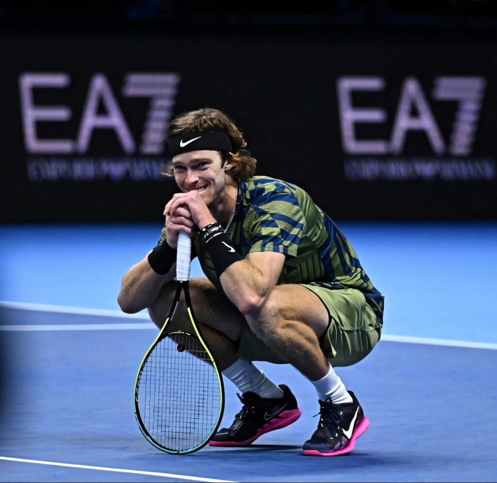 Andrey Rublev crouching on tennis court smiling holding racket wearing sporty outfit during match Wallpaper