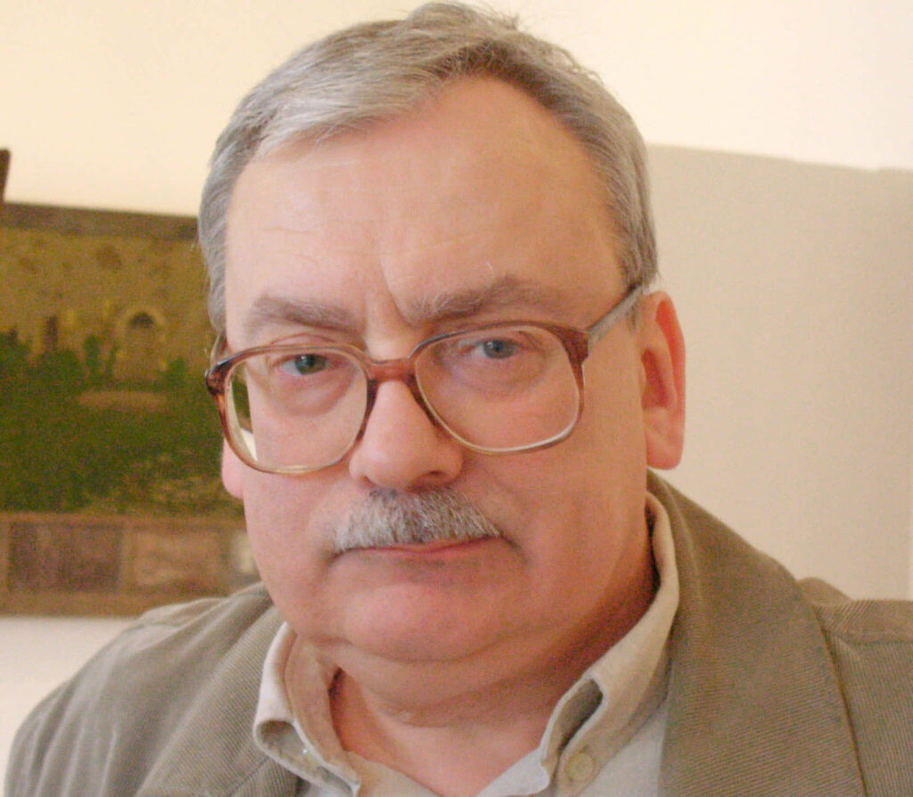 Portrait of Andrzej Sapkowski indoors with serious expression and softly lit wallpaper background