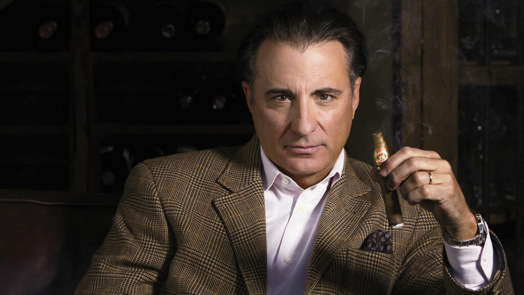 Andy Garcia stylish in checked blazer holding cigar in dim luxurious wallpaper background