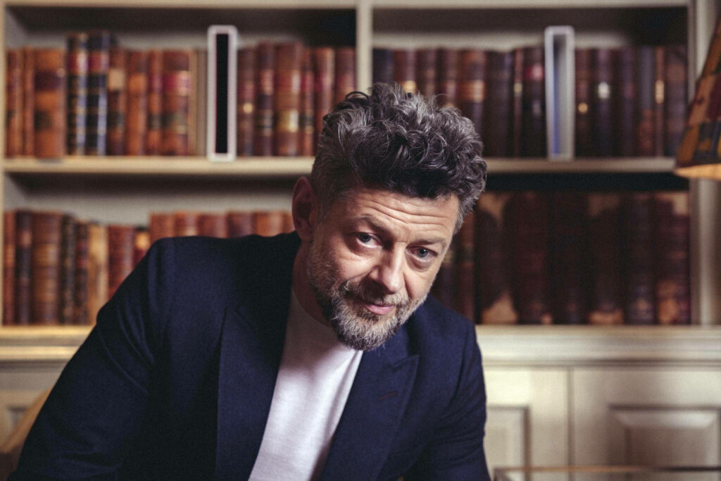 Andy Serkis seated in a cozy library with shelves of books creating a warm intellectual vibe Wallpaper