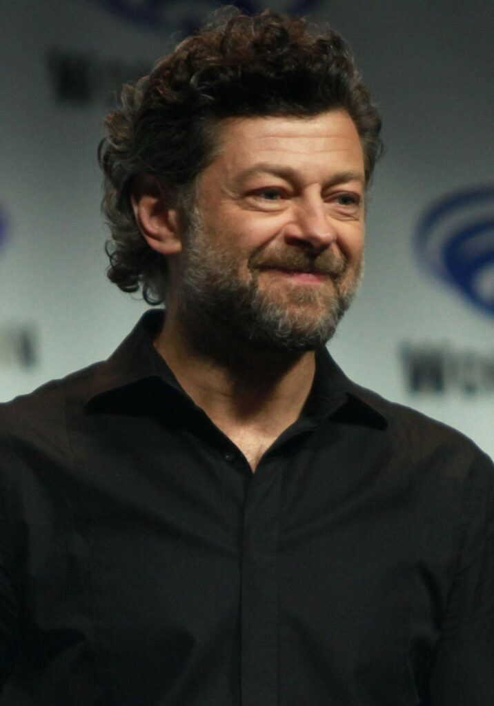 Andy Serkis smiling with rugged hair and beard in black shirt against soft wallpaper background