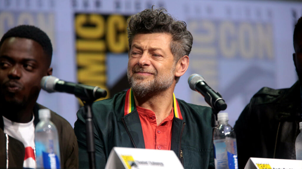 Andy Serkis smiling at Comic Con panel with microphones and wallpaper background behind him