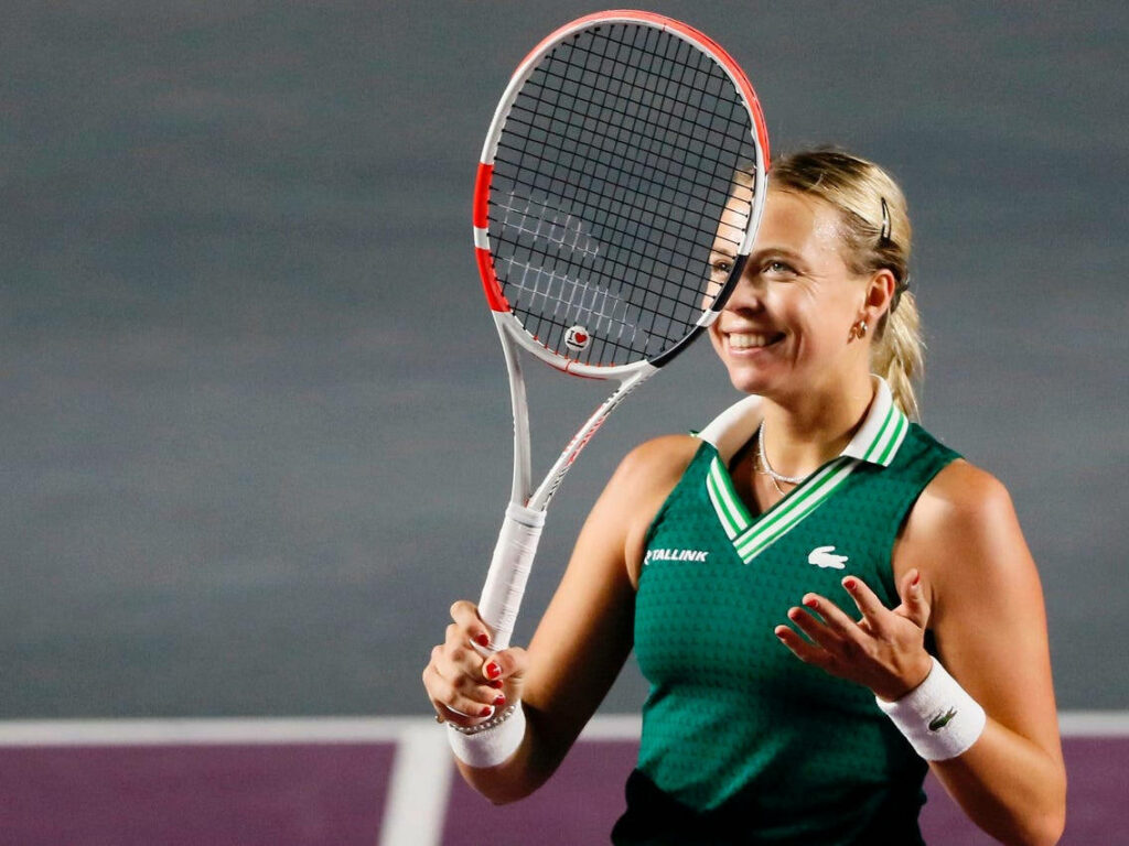 Anett Kontaveit smiling in green tennis outfit holding racket on court with wallpaper background